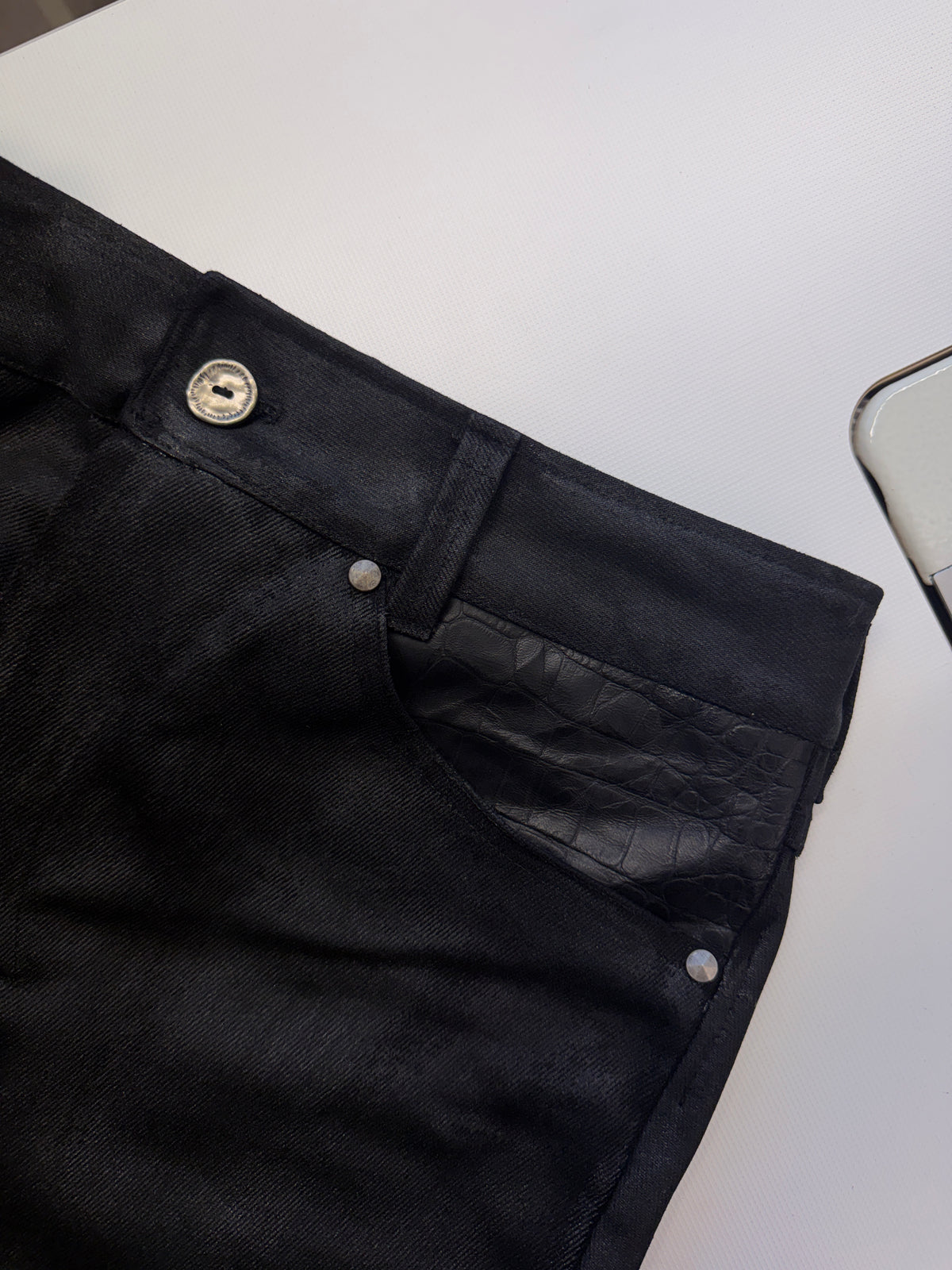 Bootcut Denim — close-up of the front pocket with black leather insert, button and rivet, laid flat on a white surface